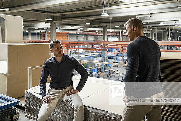 Businessman discussing with colleague while sitting on cardboard stack in industry