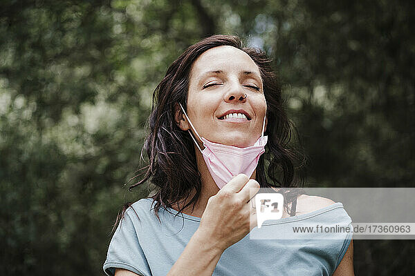 Lächelnde Frau mit geschlossenen Augen beim Abnehmen einer Schutzmaske im Wald