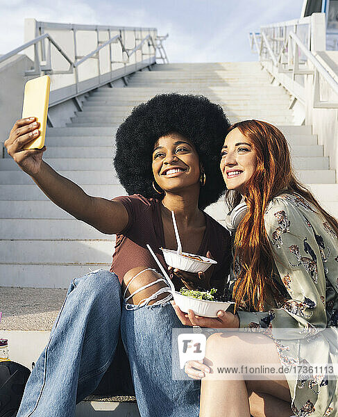 Smiling Afro woman taking selfie with female friend on steps