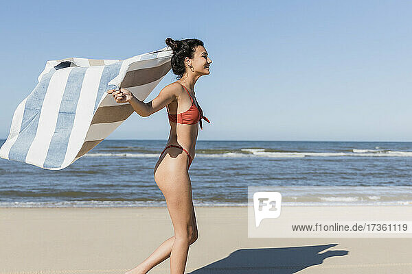 Junge Frau winkt mit dem Handtuch  während sie im Urlaub am Strand läuft