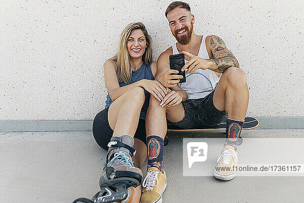 Glückliches junges Paar mit Smartphone an der Wand lehnend