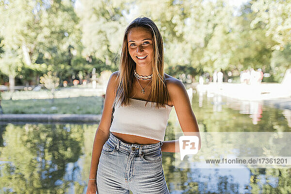 Smiling beautiful blond woman standing in front of pond at public park
