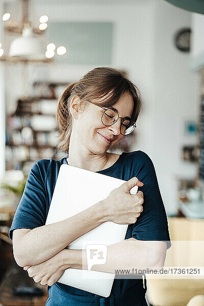 Lächelnde Freiberuflerin  die ihren Laptop umarmt  während sie in einem Cafe steht
