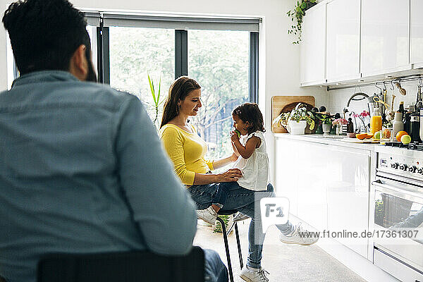 Vater sieht seine Tochter beim Obstessen an  während er mit seiner Mutter auf einem Stuhl in der Küche sitzt