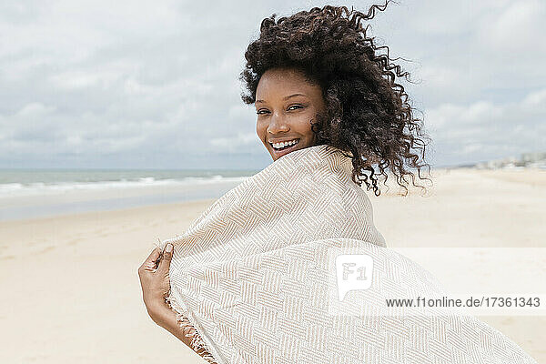 Lächelnde junge Frau mit Handtuch schaut über die Schulter am Strand