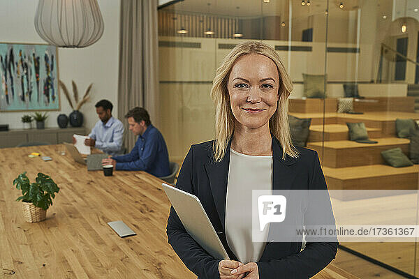 Lächelnde blonde Geschäftsfrau  die ein digitales Tablet hält  während sie im Sitzungssaal steht