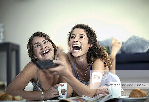 Cheerful woman changing channels with remote control while watching TV with female friend at home