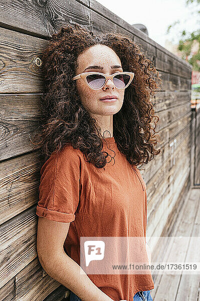 Schöne junge Frau mit lockigem Haar und Sonnenbrille lehnt an einer Holzwand