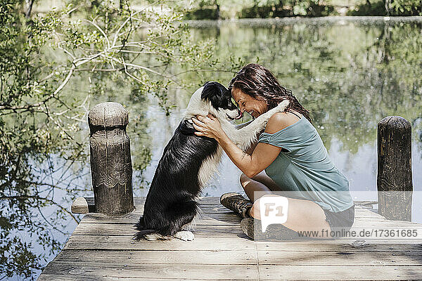 Smiling woman embracing dog while sitting on pier by lake in forest