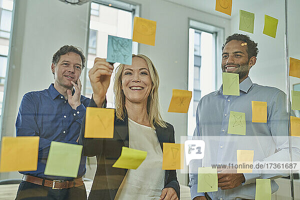 Smiling businesswoman writing on adhesive note while discussing with male colleagues in board room