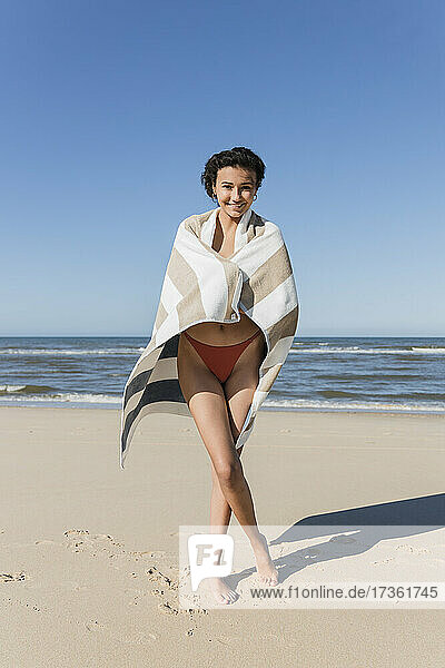 Lächelnde junge Frau  eingewickelt in ein Handtuch  steht an einem sonnigen Tag im Sand
