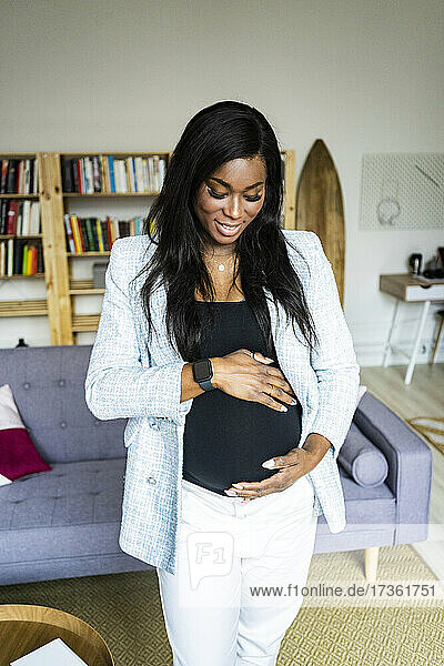 Glückliche junge schwangere Freiberuflerin  die ihren Unterleib berührt  während sie zu Hause steht
