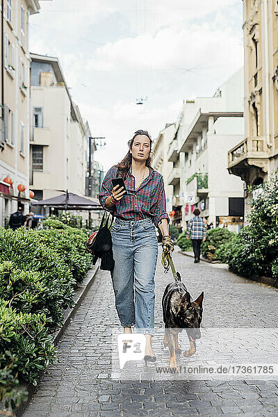 Frau hält Handy in der Hand  während sie mit ihrem Hund auf dem Gehweg spazieren geht