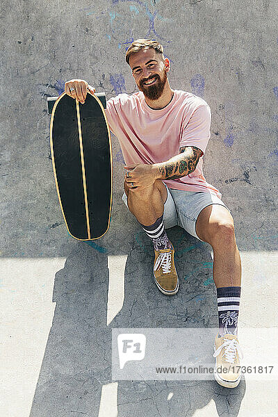 Lächelnder junger Mann sitzt mit Skateboard an einem sonnigen Tag