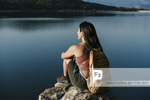 Junge Rucksackträgerin mit Rucksack auf einem Felsen am See sitzend