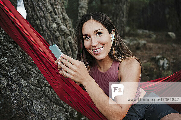 Lächelnde junge Frau mit Handy hört Musik  während sie in der Hängematte sitzt