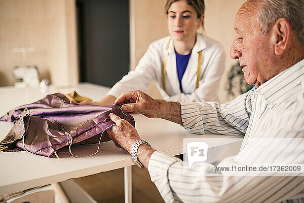 Senior male fashion expert sewing fabric while working with young female colleague
