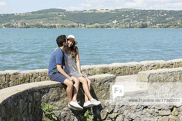 Mann küsst lächelnde Freundin  während er auf einer Stützmauer sitzt