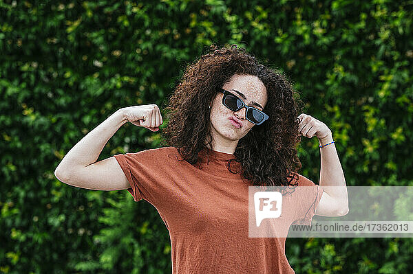 Playful young woman wearing sunglasses flexing muscles in front of ivy plants