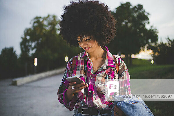 Junge Frau mit Afrofrisur  die in der Abenddämmerung im Freien ihr Smartphone benutzt