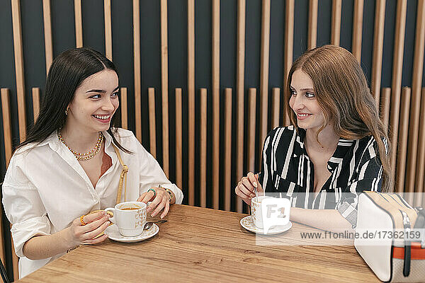 Lächelnde Freunde mit Kaffeetassen  die sich gegenseitig ansehen  während sie im Cafe sitzen
