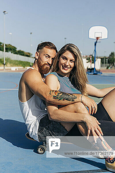 Lächelnde junge Frau sitzt mit ihrem Freund auf dem Skateboard am Basketballplatz