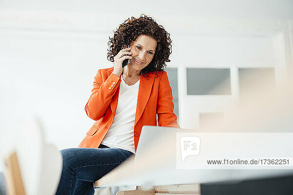 Female professional using laptop while talking on mobile phone at office
