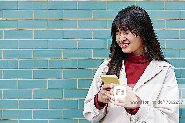 Frau mit geschlossenen Augen  die ihr Smartphone vor einer türkisfarbenen Wand hält