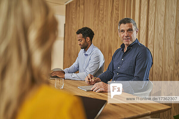 Männlicher Berufstätiger im Gespräch mit einer Mitarbeiterin  während er neben einem Geschäftsmann im Sitzungssaal sitzt