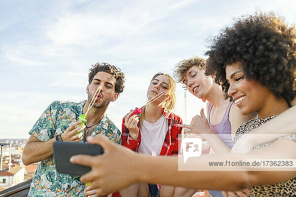 Junge männliche und weibliche Freunde blasen Seifenblasen  während sie ein Selfie auf dem Dach machen