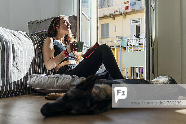 Woman with coffee cup and book relaxing by dog at home