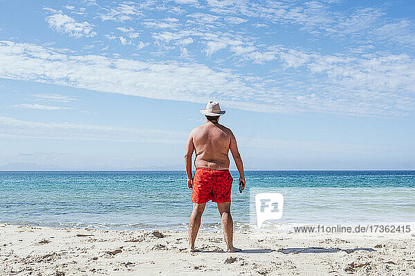 Übergewichtiger Mann mit Hut schaut an einem sonnigen Tag aufs Meer