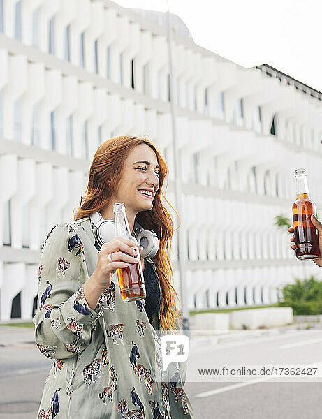 Smiling redhead woman looking at female friend while enjoying beer at roadside