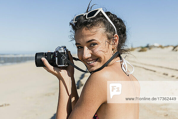 Smiling young woman with camera standing at beach on sunny day