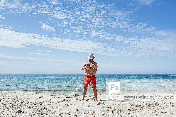 Schwuler Mann ohne Hemd benutzt Handy am Strand im Urlaub