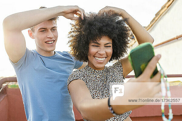 Fröhliches junges Paar  das ein Selfie mit seinem Smartphone macht  während es auf dem Dach mit seinen Haaren spielt