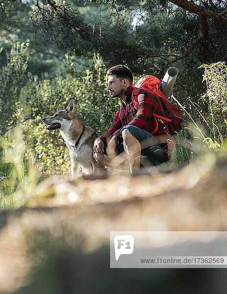 Nachdenklicher Mann mit Rucksack hockt neben einem Hund im Wald