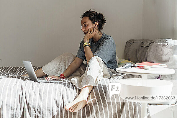Weibliche Fachkraft  die einen Laptop benutzt  während sie mit der Hand am Kinn im Schlafzimmer sitzt