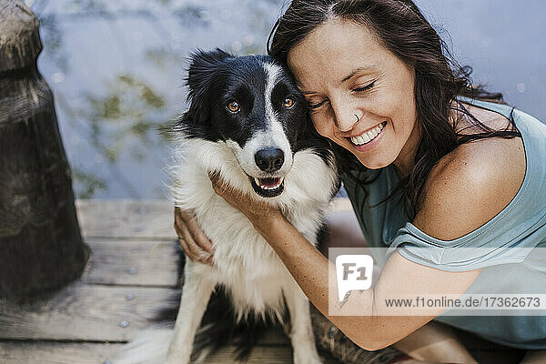 Lächelnde Frau mit geschlossenen Augen  die ihren Hund umarmt  während sie auf einem Steg sitzt