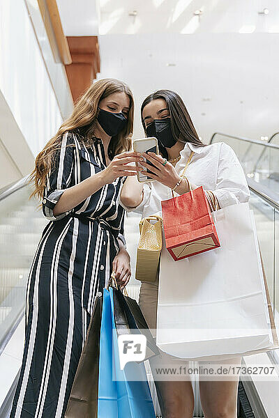 Freundinnen mit Gesichtsschutz tragen Einkaufstaschen  während sie in einem Einkaufszentrum ihr Smartphone benutzen