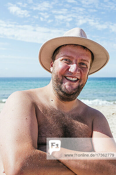 Lächelnder Mann ohne Hemd  der mit verschränkten Armen am Strand steht