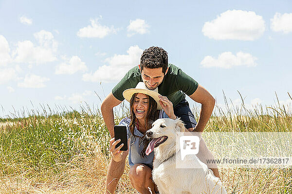 Girlfriend taking selfie with boyfriend by dog on meadow