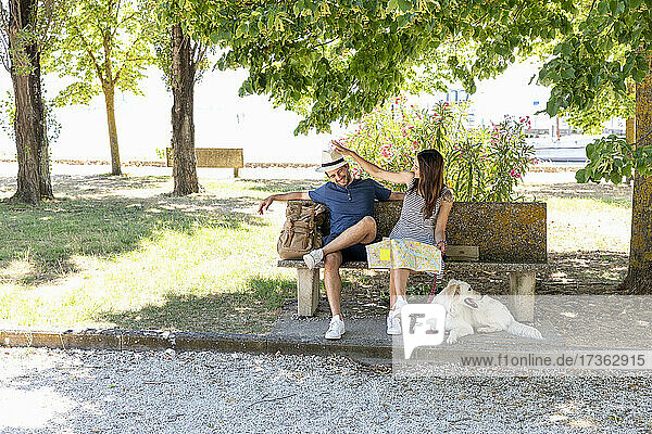 Touristenpaar mit Landkarte und Rucksack sitzt mit Hund auf einer Bank