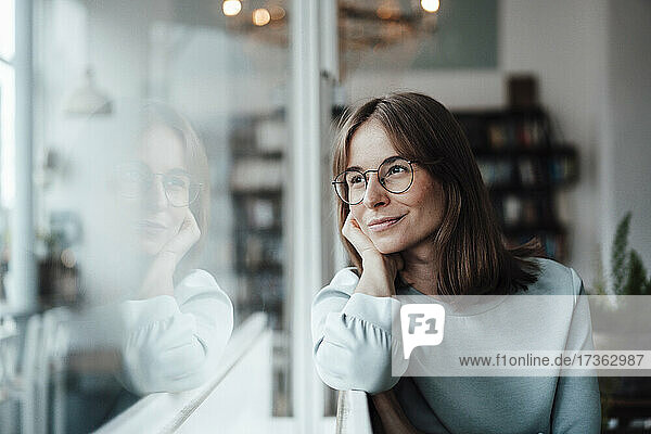 Lächelnde nachdenkliche Frau mit braunen Haaren  die durch das Fenster schaut  während sie in einem Café sitzt