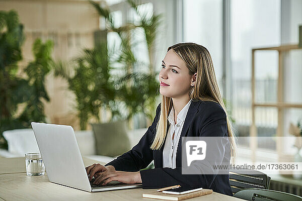 Thoughtful teenage intern looking away while sitting with laptop at desk in office