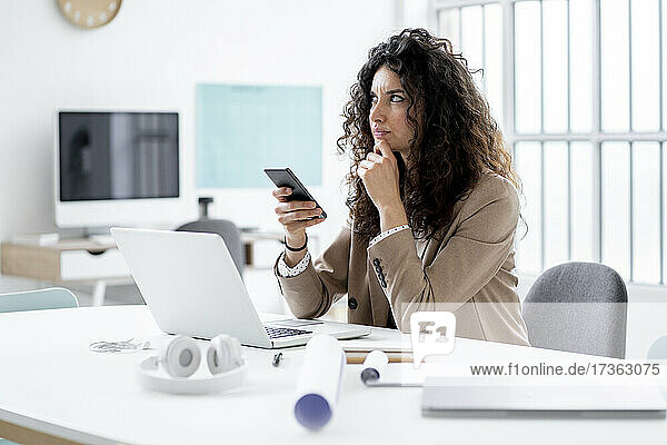 Nachdenkliche Geschäftsfrau  die aufschaut und ihr Handy am Schreibtisch hält
