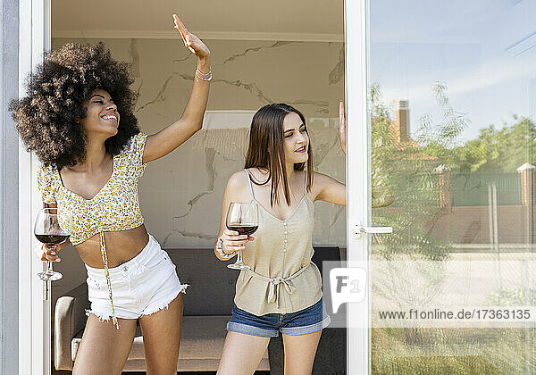 Multi-ethnische Freundinnen mit Weinglas winken  während sie an der Tür eines Hauses stehen