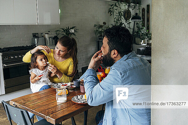 Frau füttert ihre Tochter mit Essen  während sie mit einem Mann am Tisch sitzt