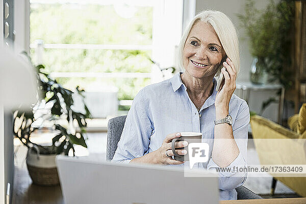Smiling businesswoman holding mug while talking on smart phone at home office
