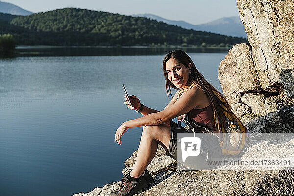 Lächelnde Wanderin mit Handy in der Hand auf einem Felsen am See sitzend